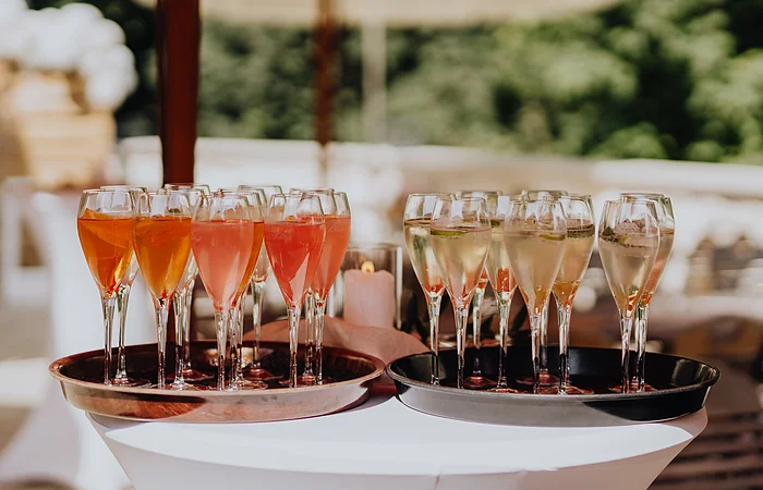 Auf einem weißen Partystechtisch befinden sich zwei braune Tabletts. Auf einem Tablet stehen Weinbläser mit weißem Sekt und auf dem anderen Weingläser mit rose farbenem Sekt.