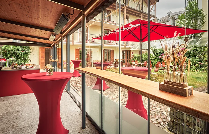 Rote Stehtische sind im Außenbereich platziert. Im Hintergrund sind weitere Sitzmöglichkeiten und ein Sonnenschirm.