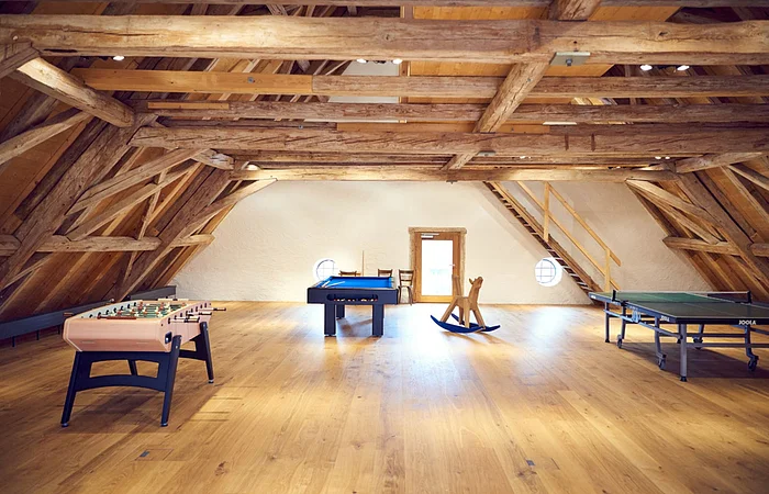 Eine große Fläche im Dachgeschoss mit Holzwänden und Deckenbalken aus Holz. Eine Tischtennisplatte und ein Kickertisch stehen im Raum.
