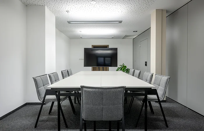 Die grauen Stühle sind um einen weißen langen Tisch mit Blick zum Bildschirm gerichtet. Der Raum ist minimalistisch eingerichtet.