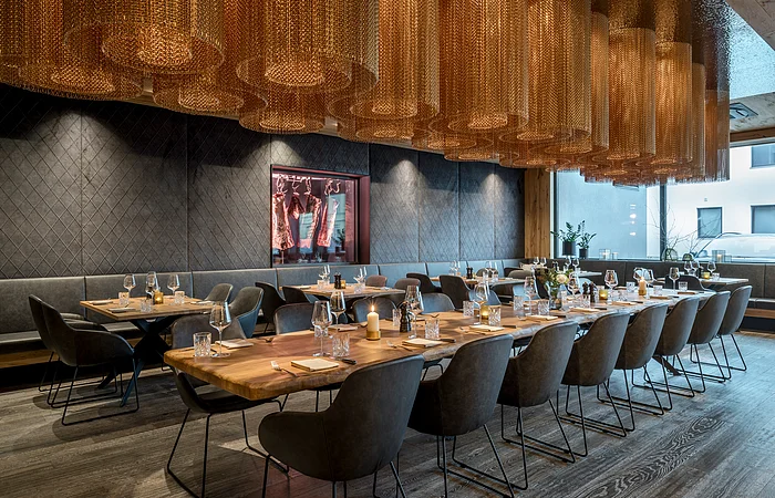 Der große und offene Raum ist mit rustikalen Holztischen und grauen Stühlen versehen. Von der Decke hängen zylinderförmige braune Lampen.