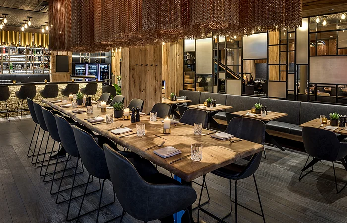 Der große und offene Raum ist mit rustikalen Holztischen und grauen Stühlen versehen, er hat eine ruhige Atmosphäre. Von der Decke hängen zylinderförmige goldene Kettenlampen.