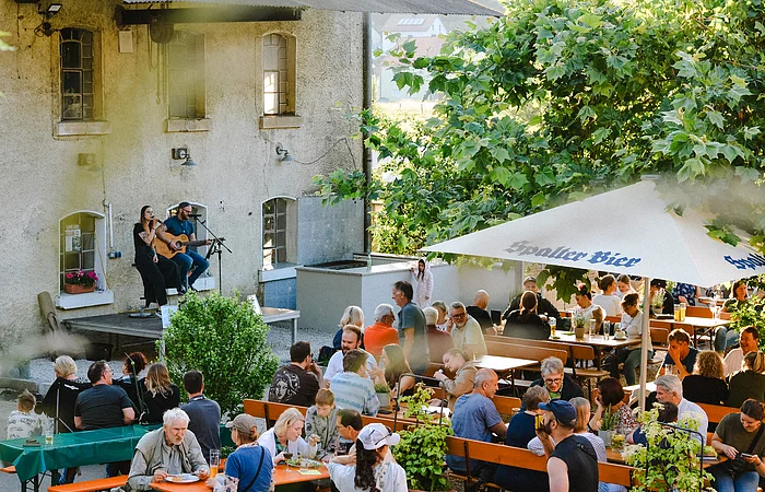 In dem Biergarten mit Bierbänke, Schirme und einer kleinen Bühne vor einem Haus der Aktiv Mühle sitzen viele Menschen gemütlich auf Ihren Plätz und hören der Musik auf der Bühne zu.