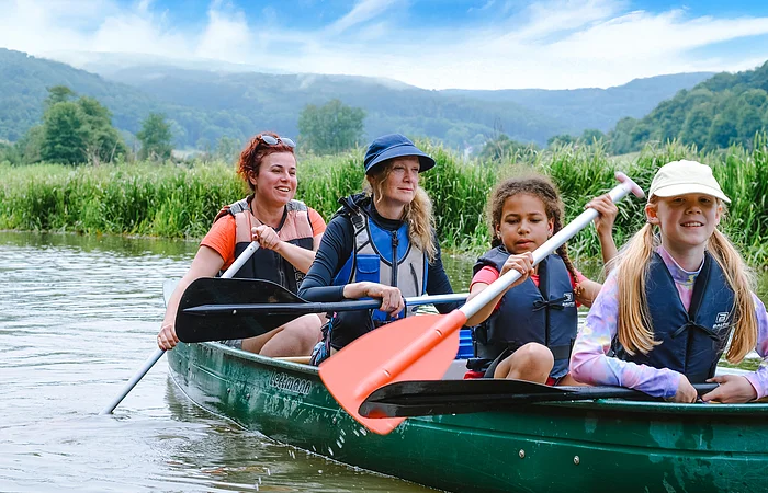 Zwei Kinder mit zwei Frauen sitzen in einem Kanu und paddeln mit Freude auf der Altmühl entlang im Hintergrund sind Wiesen und ein blauer Himmel zu sehen.