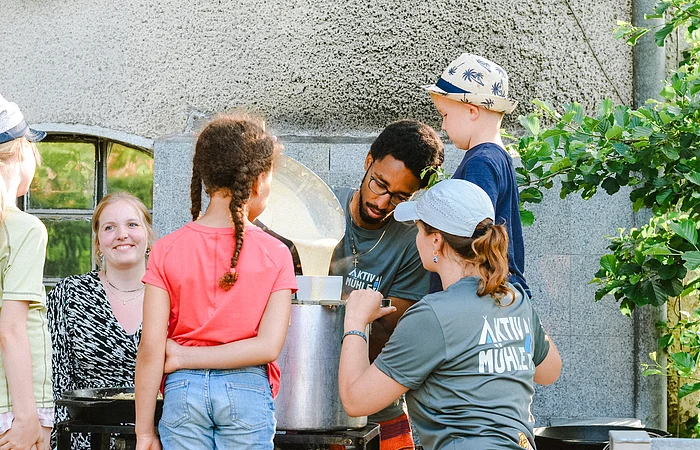 Ein Mann und eine Frau stehen mit einigen Kinder um einen großen Topf und kochen mit einander Spätzle im freien.