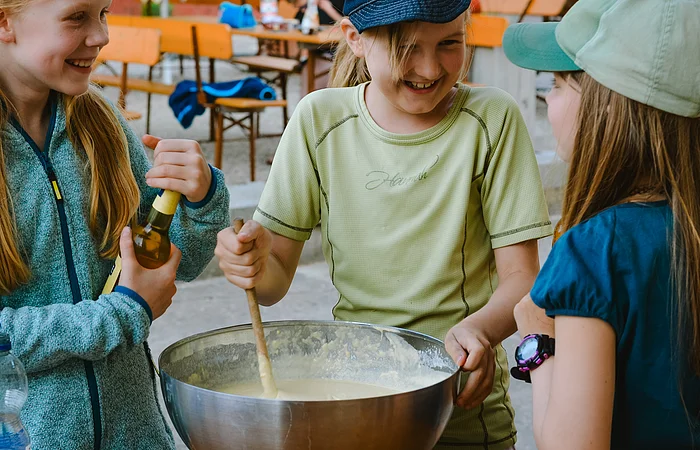 Drei Kinder stehen um eine große Schüssel voller Teig und kochen/backen miteinander im Camp der Aktiv Mühle. Die Kinder haben große Freunde und lachen über das ganze Gesicht.
