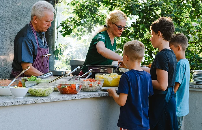 An einem Tisch mit verschiedenen Salaten und Speisen stehen 3 Kinder zum Essen holen an. Eine Frau und ein Herr geben Ihnen auf Ihr Teller die Speisen.