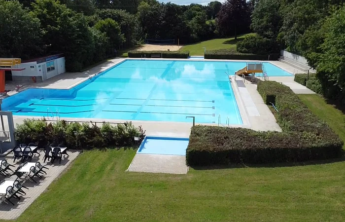 Außenansicht eines leeren Schwimmbeckens mit Sprungturm, Rutsche, Sitzbereich und umgebender Grünfläche.