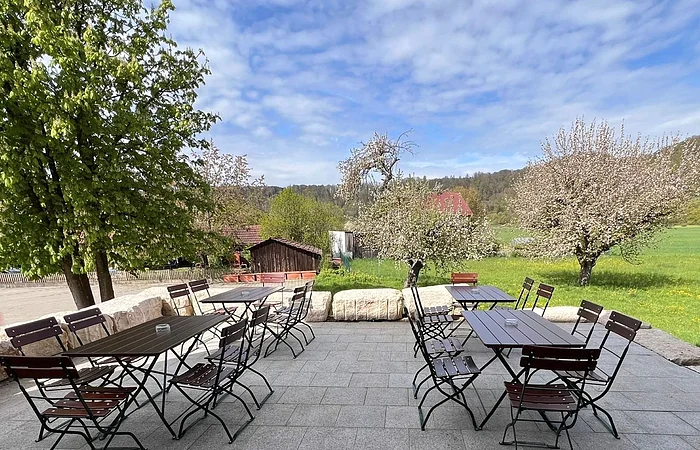 Außenterrasse mit leeren Tischen und Stühlen, umgeben von blühenden Bäumen und grüner Landschaft.