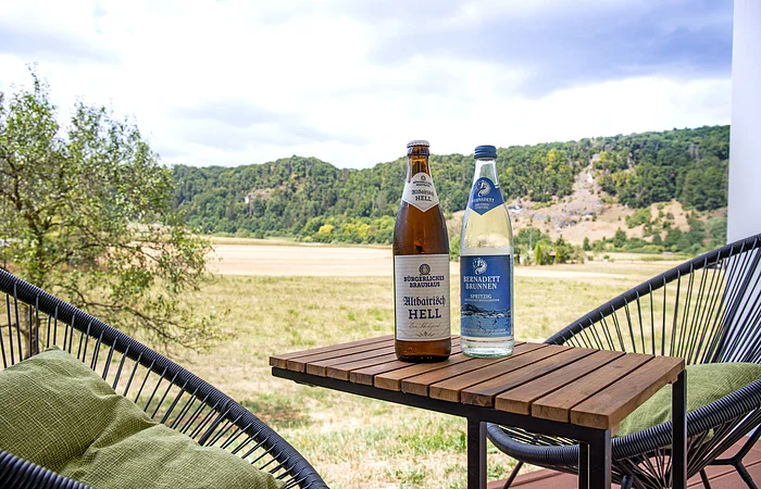Zwei Flaschen, eine Bier- und eine Wasserflasche, stehen auf einem Holztisch vor einer Landschaft mit Feldern und Bäumen.