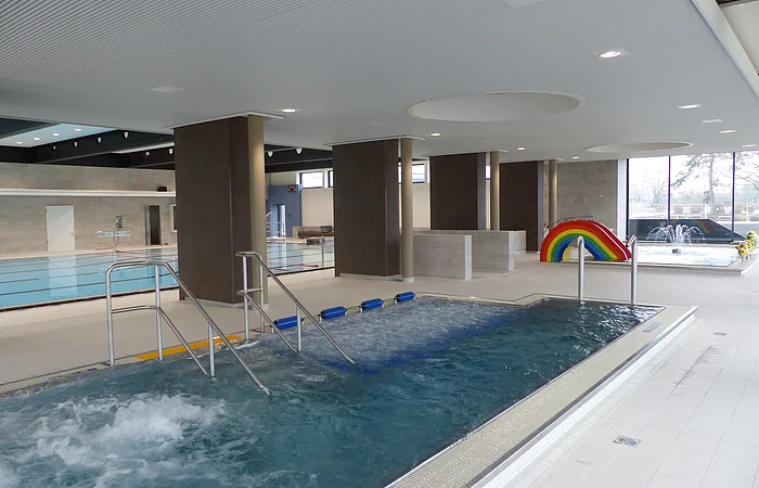 Innenansicht eines Schwimmbads mit mehreren Becken, Rutsche in Regenbogenfarben und Wasserspielen.