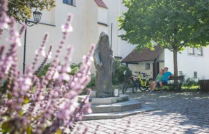 Steinfigur der heiligen Walburga auf einem gepflasterten Platz vor der Stadtpfarrkirche St. Walburga in Monheim, daneben zwei sitzende Personen auf einer Bank, dahinter ein Baum. Im Vordergrund unscharf Blumen.