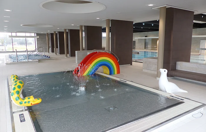 Innenansicht eines Schwimmbads, dem Jurabad Monheim, mit Kinderbecken, Regenbogenrutsche und Wasserspielzeugen. Im Hintergrund ein weiteres Schwimmbecken und ein großes offenes Fenster.