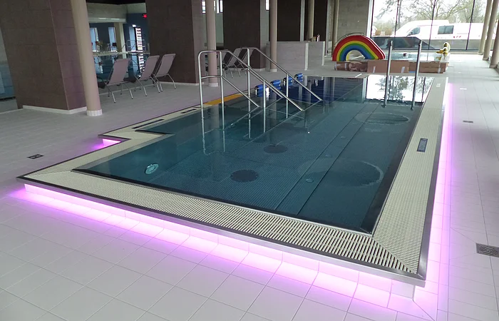 Innenansicht eines Schwimmbades, dem Monheimer Jurabad: ein mit Wasser befülltes Schwimmbecken mit Stufen und Handläufen, umgeben von Fliesenboden. Der Rand des Beckens ist außenrum rosa/lila beleuchtet. Im Hintergrund stehen drei Liegestühle und eine Regenbogenrutsche, sowie ein weiteres Becken.