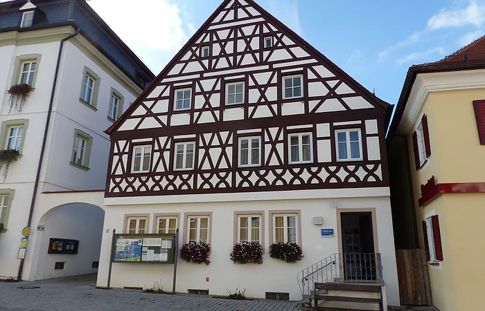 Tourist-Information der Stadt Monheim/Monheimer Alb: ein Fachwerkhaus mit drei Stockwerken, weißen Wänden und braunen Balken. Davor steht eine Sitzbank und ein Schaukasten auf einem gepflasterten Platz. Links davon ein weißes dreistöckiges Gebäude. Rechts davon ein gelbes zweistöckiges Gebäude mit roten Fensterläden.