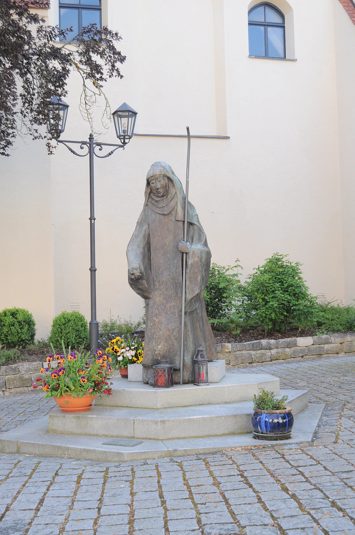 Statue der Hl. Walburga Monheim Steinstatue der heiligen Walburga in Monheim mit Stab, umgeben von Blumen in Töpfen und Laternen, vor einer hellen Gebäudewand, der Stadtpfarrkirche Monheim. Vor dem Gebäude sind Pflanzen eingesetzt.