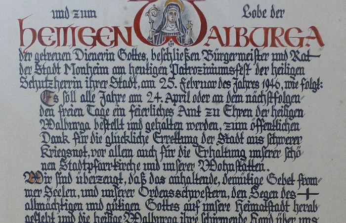 Ein handgeschriebenes Dokument mit dekorativen roten und schwarzen Buchstaben, das eine religiöse Widmung enthält. Hierin gelobt der Bürgermeister sowie der Rat der Stadt Monheim, ein alljährliches Dankamt zu Ehren der Heiligen Walburga abhalten zu lassen, für die Verschonung der Stadt im Zweiten Weltkrieg.
