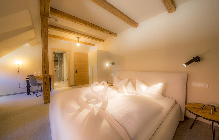 Das helle Bett steht mitten im Zimmer und ist mit Kissen verziert, der Raum hat eine Holzbalkendecke.