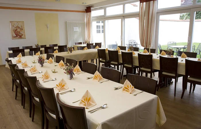 Ein großer Raum mit gedeckten Tischen, Stühlen und gefalteten Servietten, Fenster im Hintergrund.