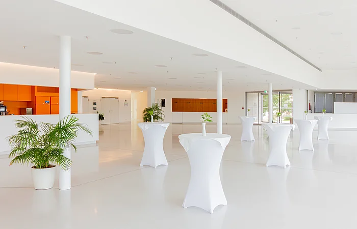 Heller Raum mit Stehtischen, weiß bezogen, Pflanzen in Töpfen, oranger Bereich links, große Fenster rechts.