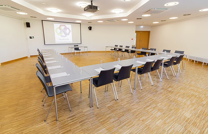 Konferenzraum mit U-förmiger Tischanordnung, Stühlen, Notizblöcken und Gläsern, Projektor und Leinwand an der Wand.