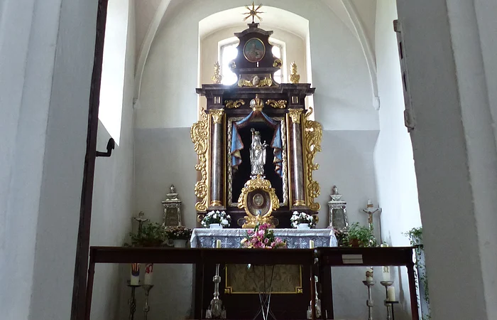 Ein Blick durch eine Tür zu einem Kirchenaltar in der Walburgakapelle in Monheim, mit goldenen Verzierungen und einer Statue, der heiligen Walburga. Der Kirchenaltar ist umgeben von Kerzen und Blumen, in einem gewölbten Raum mit einer verzierten Decke.