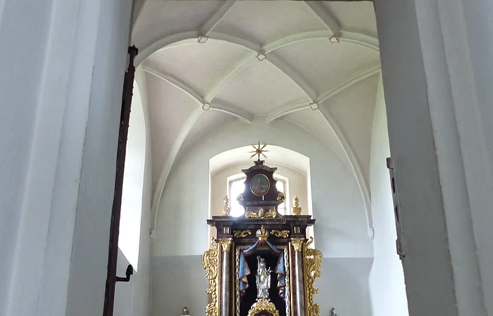Ein Blick durch eine Tür zu einem Kirchenaltar in der Walburgakapelle in Monheim, mit goldenen Verzierungen und einer Statue, der heiligen Walburga. Der Kirchenaltar ist umgeben von Kerzen und Blumen, in einem gewölbten Raum mit einer verzierten Decke.