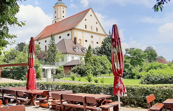 Im Vordergrund sieht man einen Teil des Biergartens mit seinen Tischen und Bänken. Im Hintergrund sieht man Wallfahrtsbasilika Maria Brünnlein.