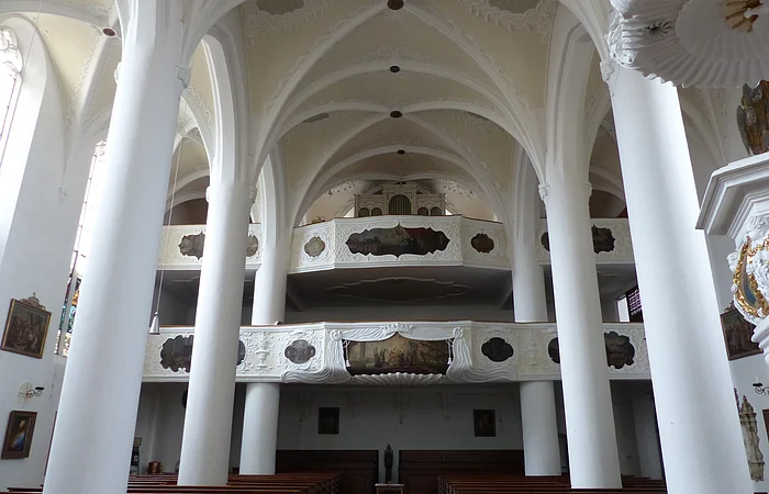 Innenansicht einer Kirche, der Stadtpfarrkirche St. Walburga in Monheim, mit hohen weißen Säulen, Gewölbedecke, Emporen mit Verzierungen und Holzbänken.