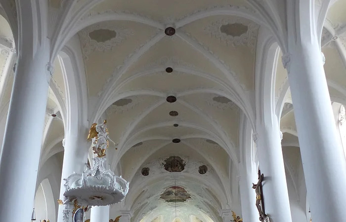 Innenansicht einer Kirche, der Stadtpfarrkirche St. Walburga in Monheim, mit hohen weißen Säulen, Gewölbedecke, Emporen mit Verzierungen und hölzernen Kirchenbänken auf beiden Seiten. Ein Altar im Hintergrund.