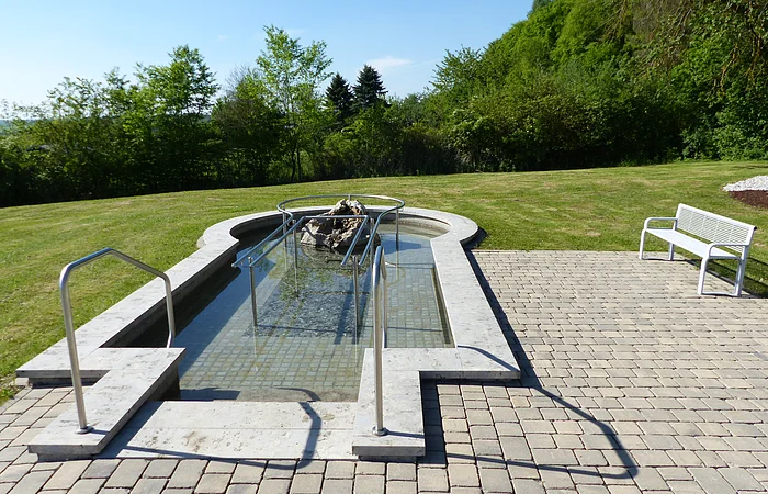 Ein befülltes Kneippbecken mit Handlauf, umgeben von Rasen und Bäumen, daneben eine weiße Bank auf Pflastersteinen.