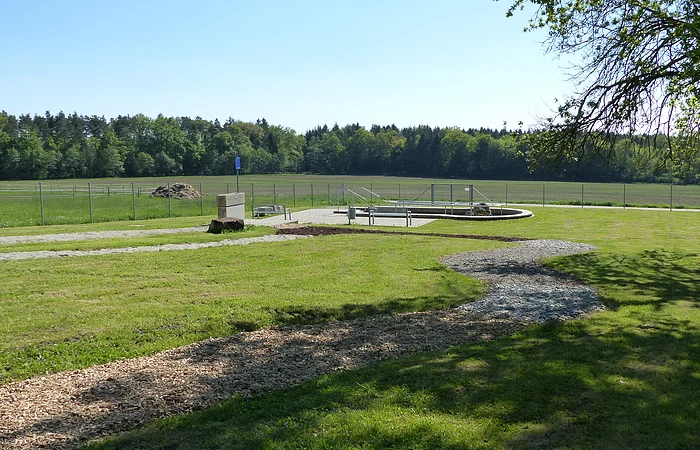 Grüne Wiese mit einem geschwungenem Barfußpfad mit Holzschnitzeln und Steinen und umzäuntem Bereich im Hintergrund, umgeben von Bäumen.