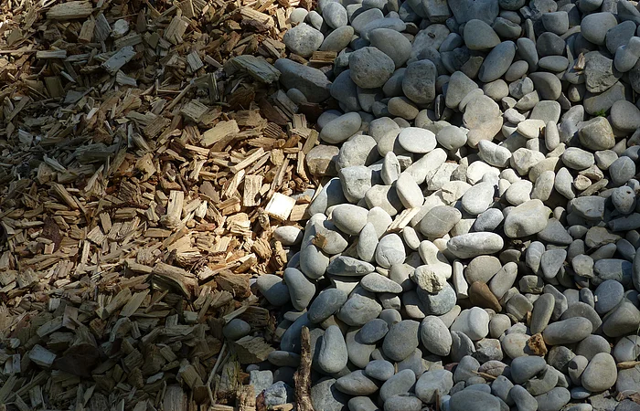 Holzhackschnitzel links neben einer Fläche mit kleinen, grauen Steinen von der Sonne bestrahlt.