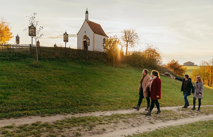 Vier Personen spazieren auf einem Weg an einer kleinen Kapelle vorbei, der Kalvarienbergkapelle in Wittesheim, umgeben von herbstlicher Landschaft. Auf dem Weg vor der Kapelle stehen Kreuzwegstationen.
