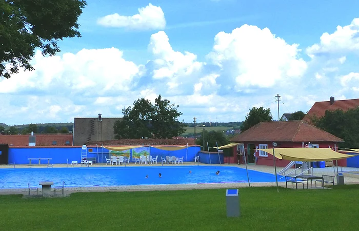 Freibad mit blauem Wasser, umgeben von Rasen, Bäumen und Gebäuden unter einem bewölkten Himmel. Vorderhalb vom Becken sind Sitzmöglichkeiten.