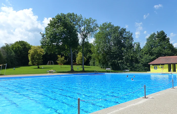 Freibad mit blauer Wasseroberfläche, im Hintergrund Bäume und ein gelbes Gebäude.