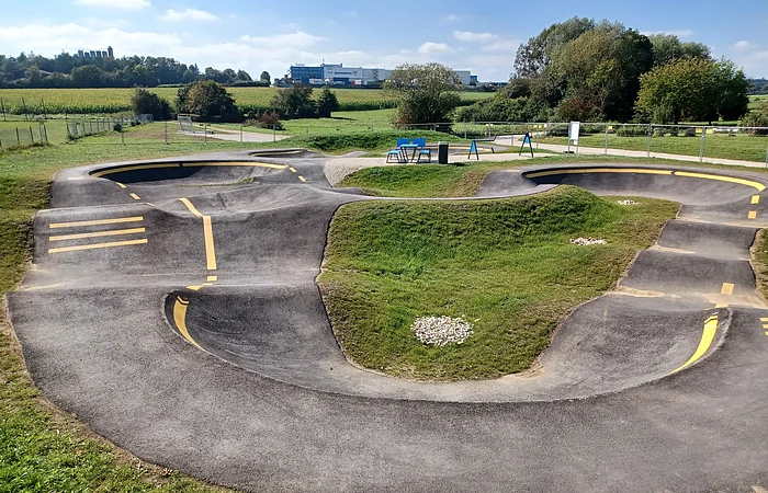 Asphaltierter Pumptrack mit gelben Markierungen in der Mitte, umgeben von grüner Landschaft und Bäumen im Hintergrund.