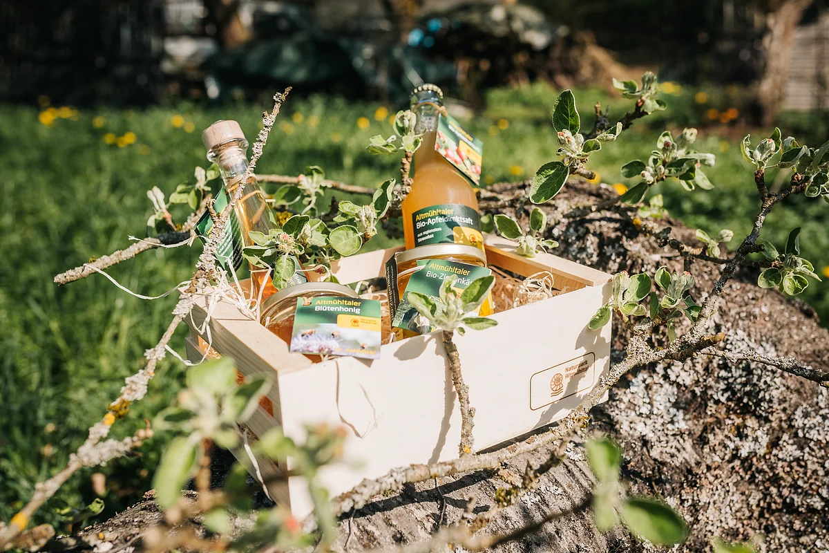 Holzkiste mit Bio-Produkten, darunter Apfelsaft, Honig und Likör, auf einem Baumstamm im Freien platziert.