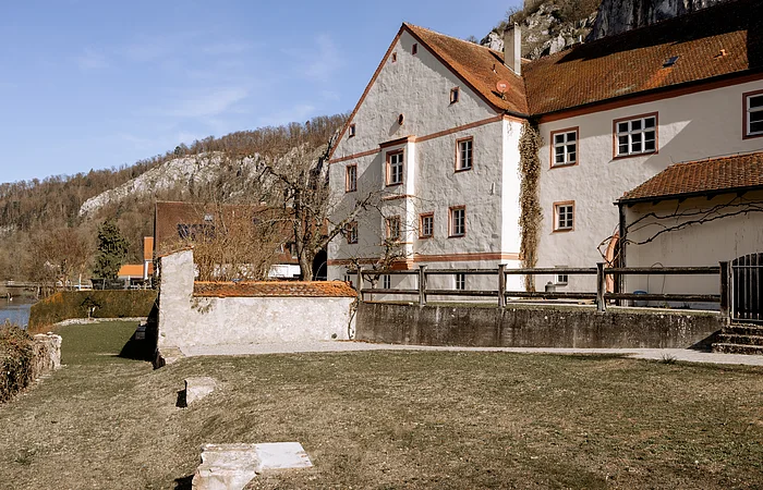 Großes, weißes Gebäude mit roten Dächern, umgeben von Bäumen und Wiese, im Hintergrund Hügel und blauer Himmel.