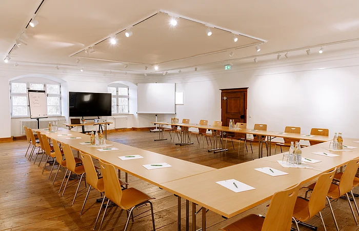 Konferenzraum mit U-förmiger Tischanordnung, Stühlen, Flipchart, großem Bildschirm und Fenstern im Hintergrund.