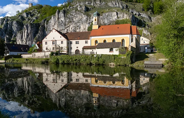 Gebäude mit rotem Dach und Turm am Flussufer, reflektiert im Wasser, umgeben von Felsen und Bäumen.