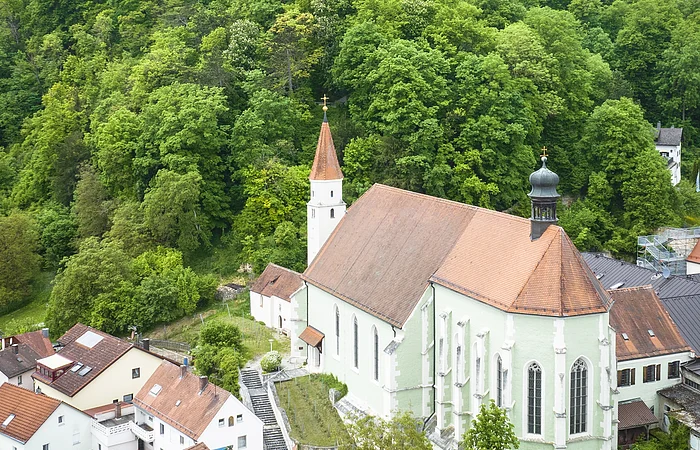 Kirche mit rotem Dach vor bewaldetem Hügel, auf dem ein rundes Gebäude steht. Wohnhäuser im Vordergrund.