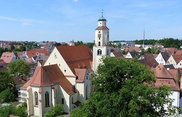 Luftbild der Stadtpfarrkirche mit Turm in einer Stadtlandschaft, umgeben von Häusern mit roten Dächern und Bäumen. Im Hintergrund blauer Himmel.