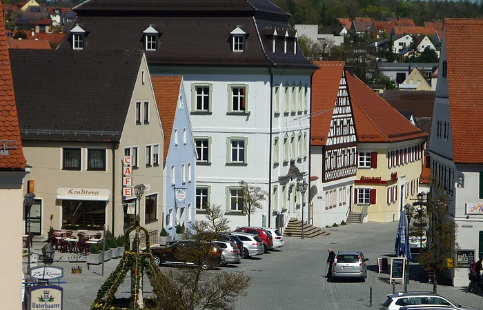 Ansicht einer gepflasterten Innenstadt mit historischen Gebäuden, geparkten Autos und einem geschmückten Oster-Brunnen. Im Hintergrund sind Wohnhäuser zu sehen.