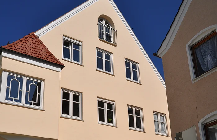 Ein dreistöckiges Gebäude mit Giebel, einem Türbogen und mehreren Fenstern auf einem gepflastertem Platz. Vor dem Gebäude steht ein Informationsschild und ein großer grauer Buchstabe "M" aus Stein. Der Buchstabe ist ein Teil des Monheimer Buchstabenweges.