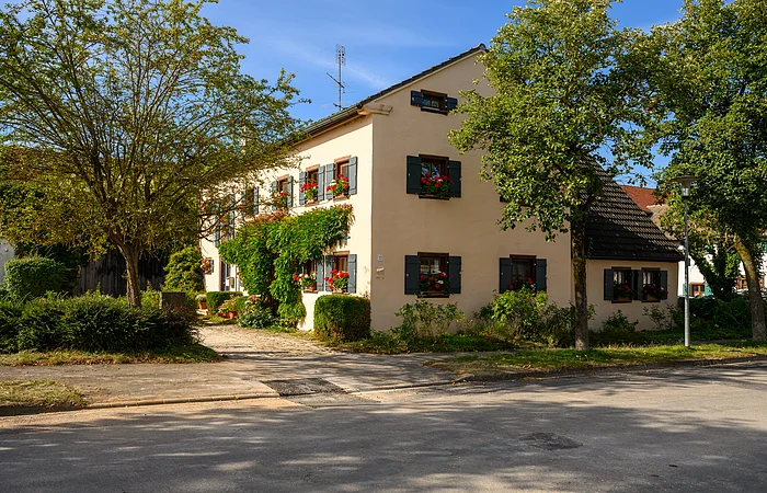 Das Familienhaus ist einen hellen Ton angestrichen und hat grüne Fensterläden, an den Fenstern befinden sich Blumenkästen mit Geranien. Um das Haus befinden sich verschiedene Sträucher und an der westlichen Häuserfront befinden sich Pflanzen
