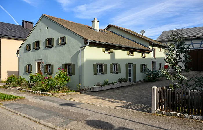 Jurahaus in Dettenheim, an der Hauptstraße gelegen, das Haus hat einen grünen Anstrich und Holzfensterläden, an der Stirnseite des Hauses befinden sich einige Sträucher