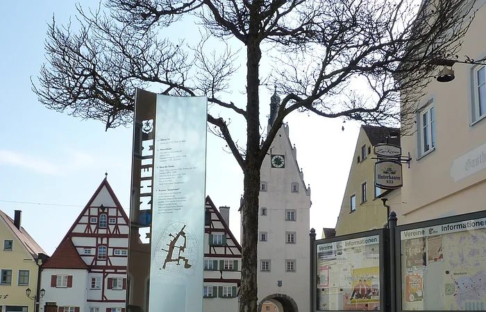 Im Vordergrund ein großer grauer Buchstabe "O" aus Stein mit einer Infotafel daneben auf einem gepflasterten Platz. Dahinter, rechts im Bild zwei weitere Informationsvitrinen der Stadt Monheim, umgeben von historischen Gebäuden im Hintergrund. Der Buchstabe ist ein Teil des Monheimer Buchstabenweges.