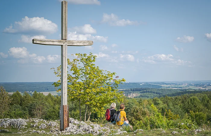 Holzkreuz auf dem Harburger Bockberg, zwei Personen mit Rucksäcken sitzen daneben, Blick auf die bewaldete Landschaft.