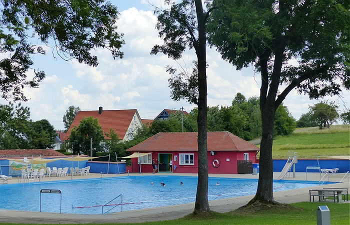 Ein Freibad mit Schwimmbecken, einer Rutsche und Sitzmöglichkeiten, umgeben von Bäumen und Wiesen. Im Hintergrund steht ein rotes Gebäude mit Fenstern, ein Kiosk.